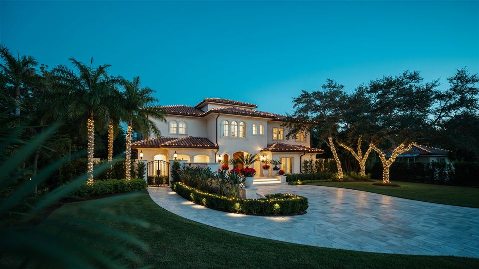 Home at twilight with lit palm trunks, wrapped oak branches and red poinsettias at the entry